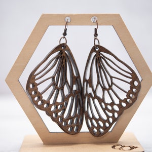 May include: A pair of wooden butterfly earrings with intricate cutouts. The earrings are brown and have a natural wood finish. They are hanging on a wooden display stand.