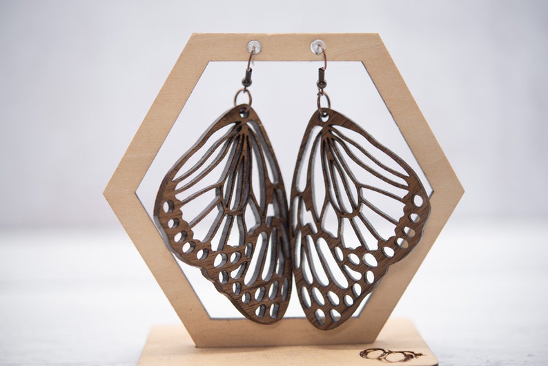 Puede incluir: Un par de pendientes de mariposa de madera con intrincados recortes. Los pendientes son marrones y tienen un acabado de madera natural. Est&aacute;n colgados de un expositor de madera.