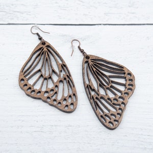 Puede incluir: Un par de pendientes de mariposa de madera con intrincados recortes. Los pendientes son marrones y tienen un aspecto delicado y natural.