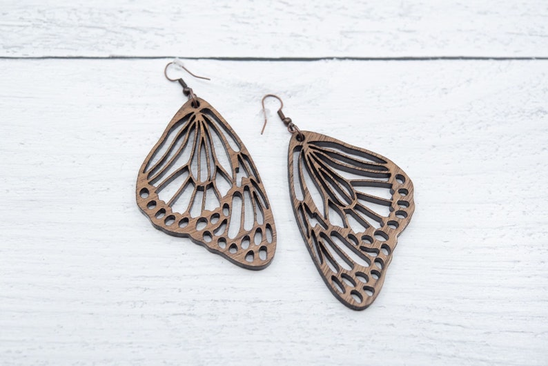 Puede incluir: Un par de pendientes de mariposa de madera con intrincados recortes. Los pendientes son marrones y tienen un aspecto delicado y natural.