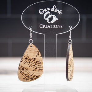 Puede incluir: Pendientes de madera en forma de lágrima con notas musicales grabadas. Los pendientes se exhiben en un soporte de acrílico transparente con el texto "Our Love Creations". Los pendientes tienen ganchos plateados.