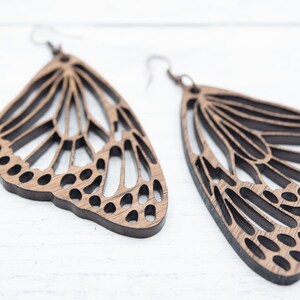 Puede incluir: Un par de pendientes de mariposa de madera con intrincados recortes. Los pendientes son de color marr&oacute;n claro y tienen un dise&ntilde;o delicado y femenino.