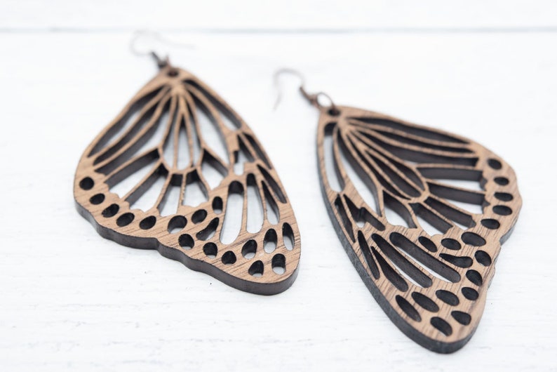 Puede incluir: Un par de pendientes de mariposa de madera con intrincados recortes. Los pendientes son de color marr&oacute;n claro y tienen un dise&ntilde;o delicado y femenino.