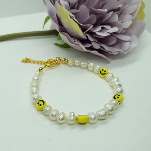 Puede incluir: Un brazalete de perlas blancas con dijes amarillos con caritas sonrientes. El brazalete tiene un cierre dorado.