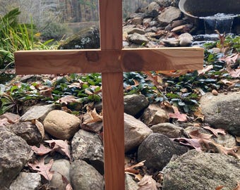 May include: A handcrafted wooden cross with a natural finish. The cross is made of light brown wood and is set against a backdrop of rocks, leaves, and a blurred natural setting. The cross is a symbol of faith and spirituality.