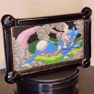 以下が含まれることがあります： 黒枠のクロスステッチ刺繍で、ピンクとグリーンのアクセントが付いた青と白の生き物が描かれている。生き物は、グレーとグリーンのパターンが施された白い背景に囲まれている。
