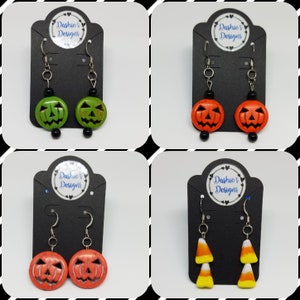 Può includere: Quattro paia di orecchini a tema Halloween. Il primo paio presenta orecchini a forma di zucca verde con perline nere. Il secondo paio presenta orecchini a forma di zucca arancione con perline nere. Il terzo paio presenta orecchini a forma di zucca arancione con perline bianche. Il quarto paio presenta orecchini a forma di mais dolce arancione e giallo con perline bianche. Tutti gli orecchini sono su schede per orecchini nere con il testo "Dashie's Designs".