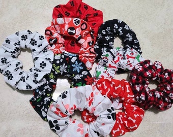 May include: A set of eight scrunchies with various dog-themed patterns. The scrunchies are made of soft, stretchy fabric and feature paw prints, bones, and snowflakes. The colors include red, white, black, and green.
