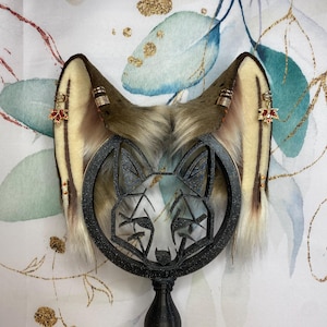 May include: A pair of faux fur animal ears in shades of brown, tan, and cream, featuring metallic gold-toned rings and small red leaf-shaped charms. The ears are mounted on a black circular stand with a geometric fox face cutout design.