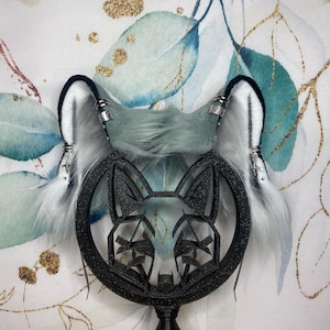 May include: A pair of gray and white faux fur fox ears with silver accents and dangling charms. The ears are attached to a black, geometric fox face centerpiece. The ears are trimmed with black and white fur.