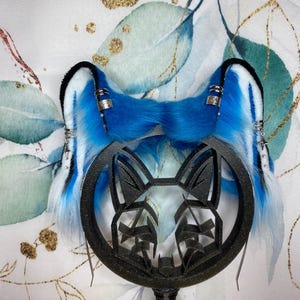 Könnte beinhalten: Ein Paar blau-weiße Kunstfellohren mit silbernen Akzenten und einem schwarzen dekorativen Rahmen. Die Ohren sind leuchtend blau mit weißen Highlights und an einem schwarzen Rahmen mit einem stilisierten Tiergesicht befestigt.