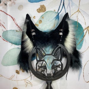 Könnte beinhalten: Schwarz-weiße Kunstfell-Katzenohren mit silbernen Akzenten. Die Ohren sind an einem schwarzen Stirnband mit einem schwarzen Katzenkopf-Ausschnitt befestigt.