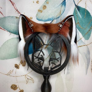 Könnte beinhalten: Fuchs-Ohren aus braunem, weißem und schwarzem Kunstfell, akzentuiert mit silberfarbenen Metallringen. Die Ohren sind auf einem schwarzen Stirnband befestigt, über einem dekorativen schwarzen Metall-Fuchsgesicht.