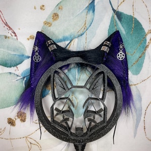 Può includere: Un copricapo decorativo con orecchie in finta pelliccia viola e nera, dettagli argentati e ciondoli a pentagramma. Un disegno geometrico di una faccia di volpe è centrato su una cornice circolare nera.