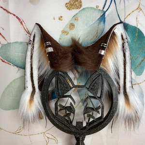 Könnte beinhalten: Ein Paar braune und weiße Kunstfell-Fuchsohren mit silbernen Akzenten. Die Ohren sind an einer schwarzen, geometrischen Fuchskopfbasis befestigt. Der Hintergrund zeigt ein Blumenmuster mit goldenen Akzenten.