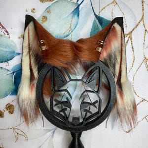 May include: A pair of fox-themed furry ear headbands with a black fox silhouette on a stand. The earbands are brown, white and red with gold accents.