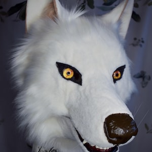 Op de afbeelding: Een wit wolvenmasker met lang imitatiebont, puntige oren en doordringende amberkleurige ogen met zwarte markeringen. De snuit heeft een donkerbruine neus en een open mond die witte tanden laat zien. De handtekening Xivafox is linksonder zichtbaar.