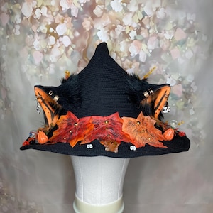Könnte beinhalten: Ein schwarzer, gehäkelter Spitzhut mit zwei Kunstfell-Tierohren, die mit kleinen Metallringen und Anhängern verziert sind. Die Krempe ist mit orangefarbenen und roten Stoff-Ahornblättern, Miniatur-Kürbissen und kleinen Wackelaugen geschmückt.