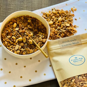 Könnte beinhalten: Eine weiße Schüssel gefüllt mit Müsli, ein goldener Löffel und ein kleiner Haufen Müsli auf einem weißen Tuch mit goldenen Punkten. Ein Kraftpapierbeutel mit dem Text "Crunchy Queen" und "Eiwess-Quelle Crunchy Müsli" ist ebenfalls zu sehen.