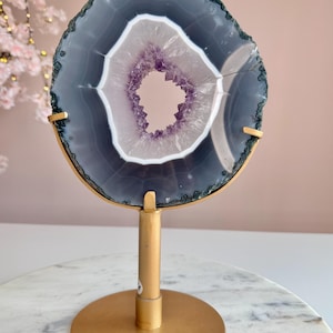 May include: A circular agate slice featuring a dark gray outer ring, a white band, and a central hollow filled with purple amethyst crystals. The stone is mounted on a gold-colored metal stand with a circular base and three prongs.