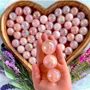 May include: A collection of iridescent, pale pink spherical stones arranged in a heart-shaped wooden bowl, with three additional matching spheres held in a palm. The stones have a smooth, reflective surface with a soft, pearlescent sheen.