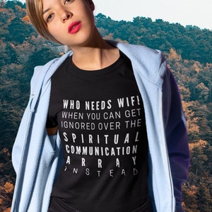 Puede incluir: Una mujer con una camiseta negra con el texto "WHO NEEDS WIFI! WHEN YOU CAN GET IGNORED OVER THE SPIRITUAL COMMUNICATION ARRAY INSTEAD" y una sudadera con capucha azul claro. Está de pie frente a un fondo de árboles.