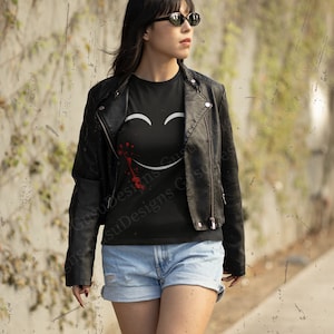 以下が含まれることがあります： 黒いTシャツを着た女性。Tシャツには白いスマイリーフェイスのグラフィックと赤いしぶきが描かれている。Tシャツの左下には「Gusu Designs」の文字が書かれている。彼女は黒い革のジャケットと青いデニムのショートパンツを着用している。