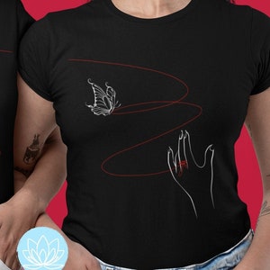 Può includere: Due magliette nere con un designo a linea bianca che presenta una farfalla, un fiore e mani con anelli rossi. Le magliette sono indossate da due persone in piedi una accanto all'altra su uno sfondo rosso.