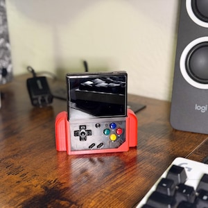 Pode incluir: Um console de jogos portátil vermelho com tela preta e botões coloridos. O console possui um direcional, botões de ação e uma tela retangular. O dispositivo está em uma superfície de madeira.