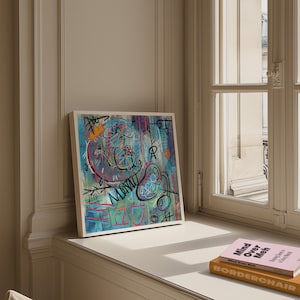 Op de afbeelding: Vierkante ingelijste abstracte kunstprint met een witte rand. Het kunstwerk toont een mix van kleuren, waaronder blauw, roze en geel, met gekrabbelde lijnen en vormen. Het woord "MONU" is zichtbaar.