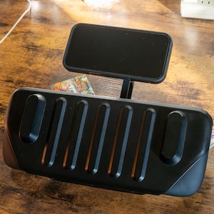 May include: A black phone stand featuring a rectangular base with vertical grooves and a phone holder. The phone holder is angled upwards, displaying a black phone. The stand is designed for desk use.