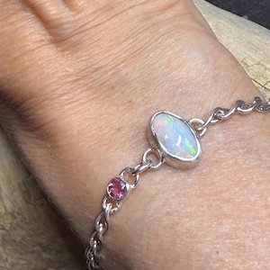Op de afbeelding: Een zilveren kettingarmband met een centrale ovaalvormige witte opaal met iriserende kleurflitsen en een kleine, ronde, roze edelsteen gezet in een zilveren zetting, verbonden door gepolijste zilveren schakels voor een verfijnd sieraad.