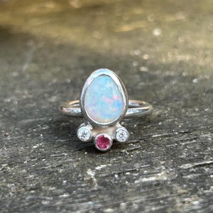 May include: A silver ring featuring a large, pear-shaped iridescent opal as the central stone. Below the opal, the band is accented with three smaller, round-cut stones: a central pink gemstone flanked by two clear, colorless stones in silver settings.
