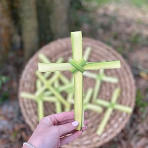 May include: A handmade cross crafted from fresh green palm leaves, held in a hand. Several similar crosses are visible in a woven basket in the background, with a blurred outdoor setting.