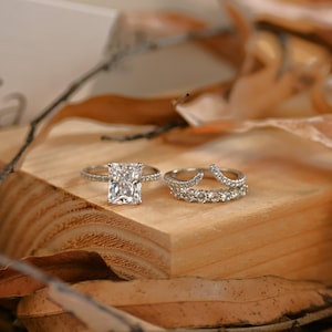 May include: A silver-toned engagement ring featuring a large rectangular radiant-cut center stone and a pavé band, paired with a matching curved wedding band set with multiple small round stones along the top edge.
