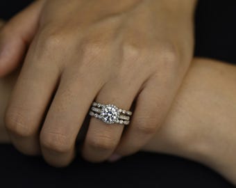 Könnte beinhalten: Ein silberfarbenes Ring-Set bestehend aus drei gestapelten Ringen. Der mittlere Ring hat einen großen runden Brillantschliff-Stein mit einem Kranz aus kleineren Steinen, flankiert von zwei passenden Ringen mit Perlen- oder Pavé-Struktur.