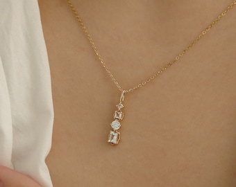 May include: A gold chain necklace featuring a vertical pendant with four clear, faceted stones in varying shapes, including a small round stone, a square stone, a larger round stone, and a rectangular emerald-cut stone at the base.