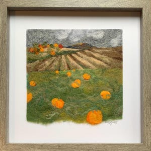 May include: A framed felt artwork showing a harvest scene. The image includes a field of pumpkins, a harvested field, and a cloudy sky. The frame is made of light grey wood.