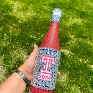 May include: A red glitter wine bottle decorated with silver rhinestones and a red and white college logo with the year 2024.
