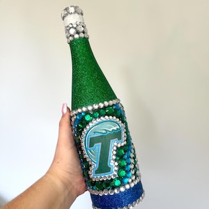 May include: A green glitter-covered bottle with a blue and green design, embellished with rhinestones. The bottle features a large letter "T" in a blue and green design. The bottle's top is adorned with rhinestones.