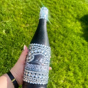 K&ouml;nnte beinhalten: Eine schwarze Flasche, die mit glitzernden Strasssteinen und einem silbernen Folienverschluss verziert ist. Die Flasche tr&auml;gt den Text "Providence Friars" in Wei&szlig; auf schwarzem Hintergrund. Die Flasche ist mit klaren, runden Strasssteinen verziert.