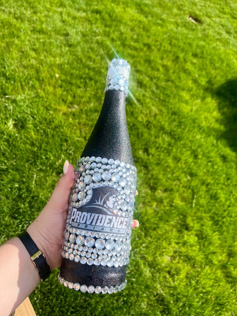 K&ouml;nnte beinhalten: Eine schwarze Flasche, die mit glitzernden Strasssteinen und einem silbernen Folienverschluss verziert ist. Die Flasche tr&auml;gt den Text "Providence Friars" in Wei&szlig; auf schwarzem Hintergrund. Die Flasche ist mit klaren, runden Strasssteinen verziert.