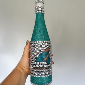 K&ouml;nnte beinhalten: Eine t&uuml;rkis glitzernde Flasche, verziert mit klaren Strasssteinen und dem Logo der Coastal Carolina University. Hals und Boden der Flasche sind mit Strasssteinen verziert, was ein dekoratives und auff&auml;lliges Design ergibt.