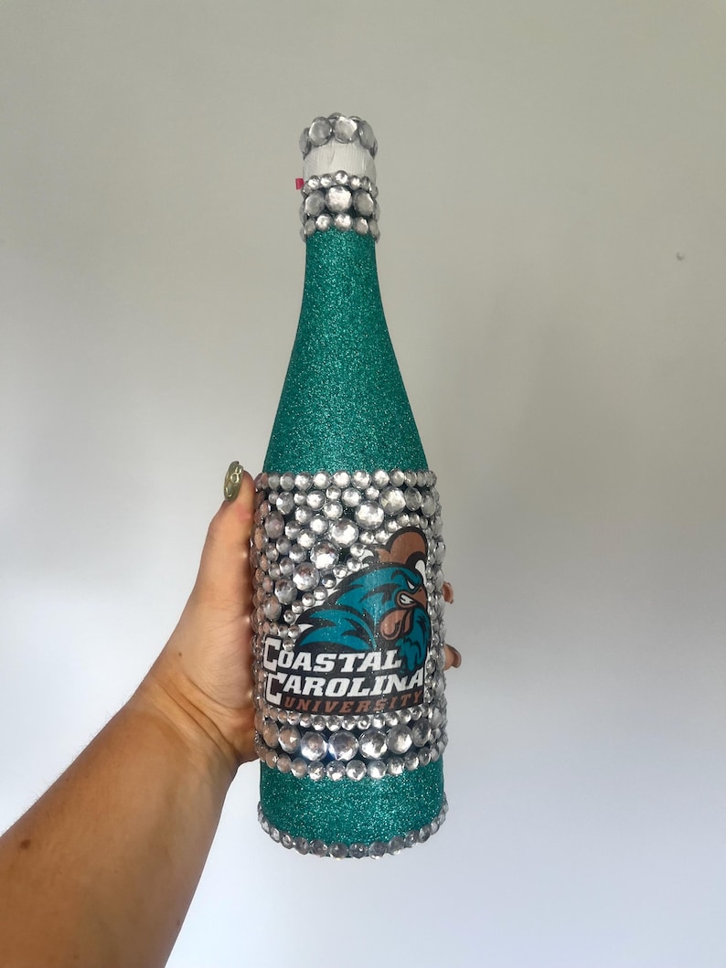 K&ouml;nnte beinhalten: Eine t&uuml;rkis glitzernde Flasche, verziert mit klaren Strasssteinen und dem Logo der Coastal Carolina University. Hals und Boden der Flasche sind mit Strasssteinen verziert, was ein dekoratives und auff&auml;lliges Design ergibt.
