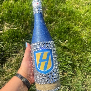 K&ouml;nnte beinhalten: Eine dekorierte Champagnerflasche mit blauem Glitzer-Finish und klaren Kristallverzierungen. Die Flasche hat ein gold-blaues Emblem mit dem Buchstaben "H" und einen goldglitzernden Sockel.