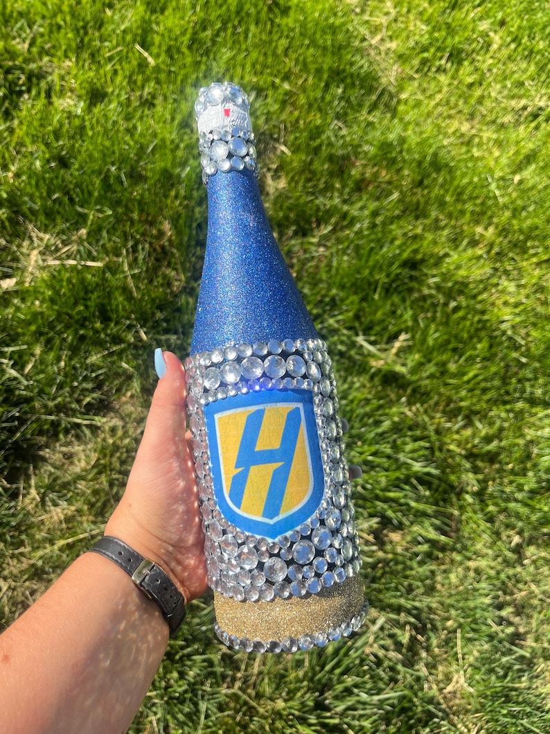 K&ouml;nnte beinhalten: Eine dekorierte Champagnerflasche mit blauem Glitzer-Finish und klaren Kristallverzierungen. Die Flasche hat ein gold-blaues Emblem mit dem Buchstaben "H" und einen goldglitzernden Sockel.
