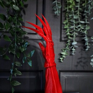 May include: A 3D printed red hand with long, sharp claws. The hand is attached to a red base.