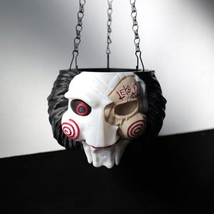 May include: Hanging planter shaped like a skull with black hair and a white and tan face. The face has red spiral designs on the cheeks and a red eye. The planter is suspended by a black chain.