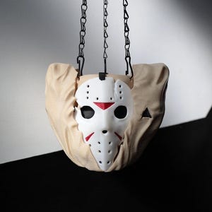 May include: A white hockey mask with red detailing and black eye holes, suspended from a black chain. The mask is set against a tan fabric backdrop, with a black and white background.