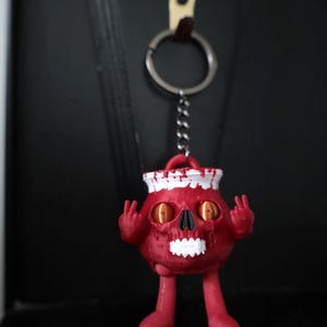 May include: A red skull-shaped keyring with a white sailor hat and peace sign hand gestures. The keyring has a silver ring and chain. The skull has orange eyes and a black nose. The keyring is hanging against a dark background.